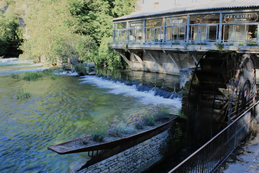 Historic wooden waterwheel and stone boat planter filled with flowers on the emerald green Sorgue River in Fontaine-de-Vaucluse, Provence.
