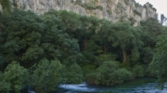 Long exposure landscape photography of the massive limestone cliffs and dense woodland canyon overlooking the emerald waters of the Sorgue river in Fontaine-de-Vaucluse, France.