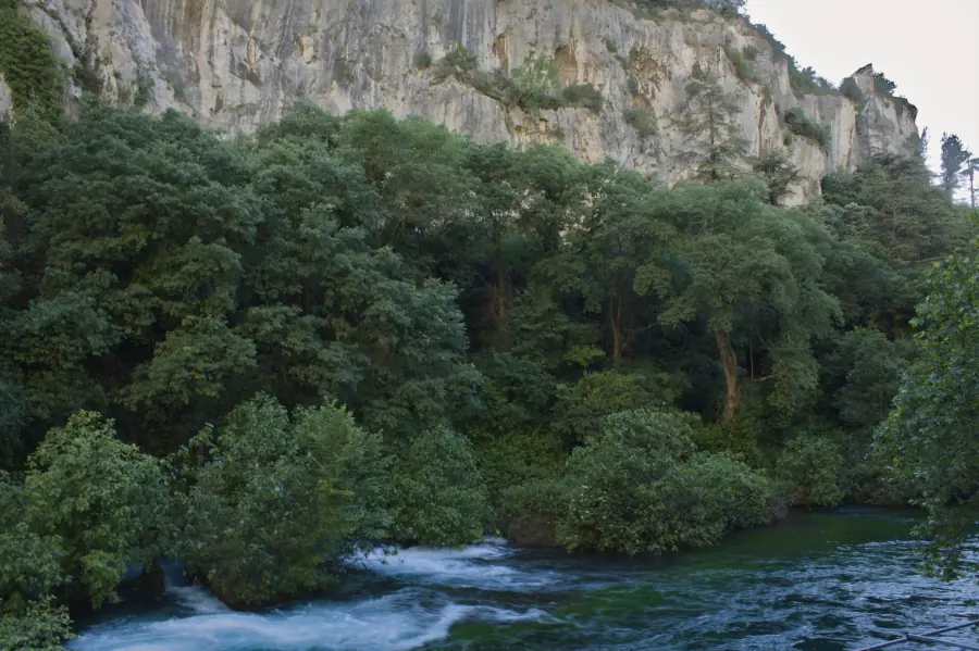 Long exposure landscape photography of the massive limestone cliffs and dense woodland canyon overlooking the emerald waters of the Sorgue river in Fontaine-de-Vaucluse, France.