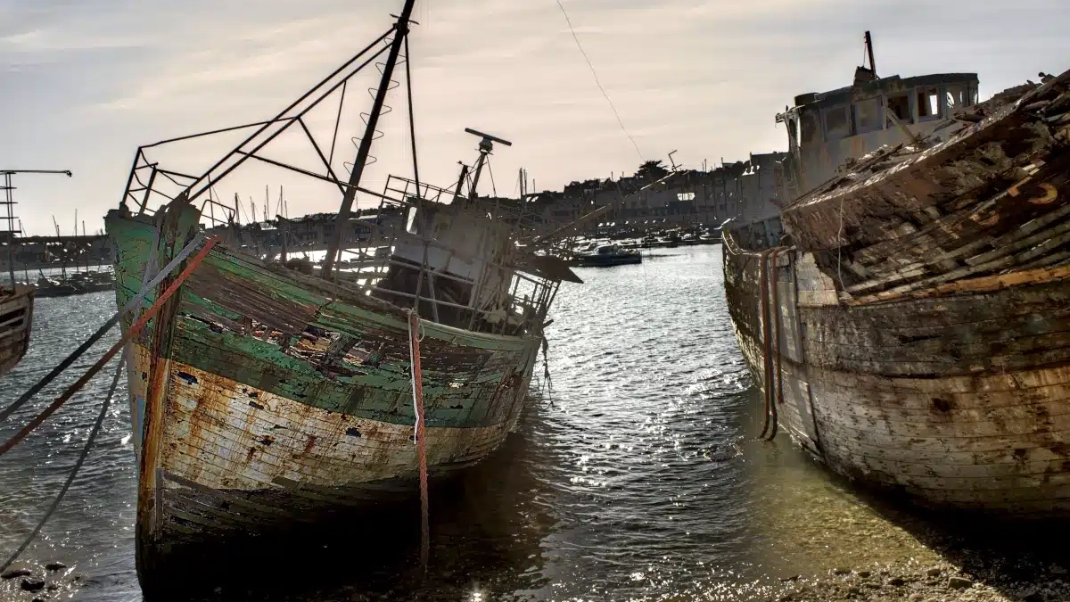 Two weathered wooden shipwrecks at the Camaret-sur-Mer boat graveyard, resting on a pebble beach at low tide under a hazy sky