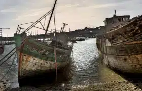 Two weathered wooden shipwrecks at the Camaret-sur-Mer boat graveyard, resting on a pebble beach at low tide under a hazy sky