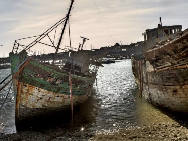 Two weathered wooden shipwrecks at the Camaret-sur-Mer boat graveyard, resting on a pebble beach at low tide under a hazy sky