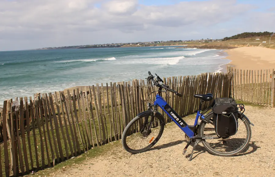 A blue T-Bird electric bicycle with travel panniers parked on a coastal path overlooking a sandy beach and the ocean.